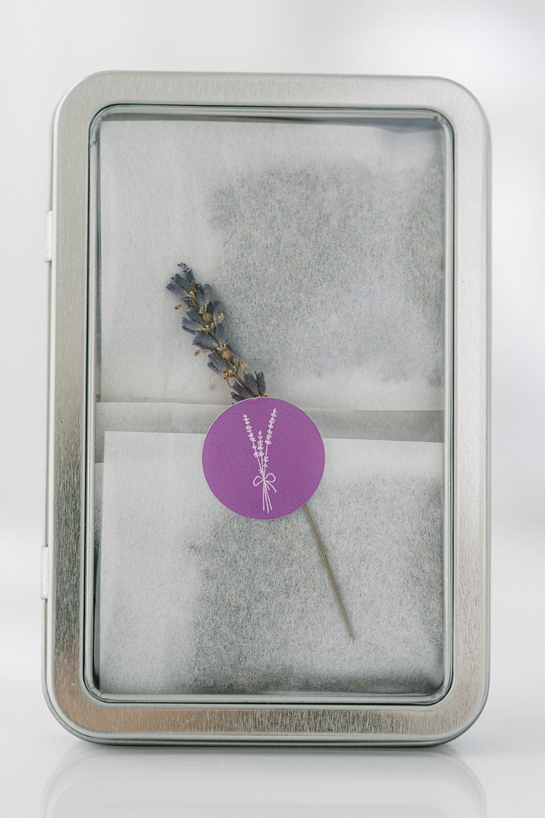 Dried Lavender with a purple label in a silver tin on a white background. Inside are lavender sachets.