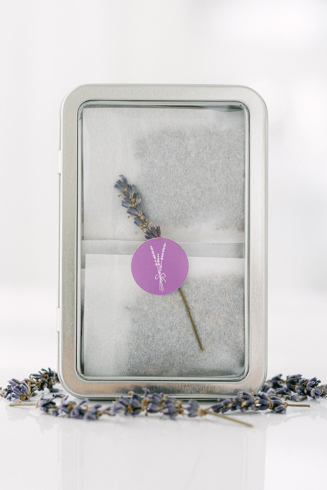 Silver tin with lavender flower and a purple label on a white background surrounded by dried lavender indicating the contents of the lavender sachets contained inside the tin.