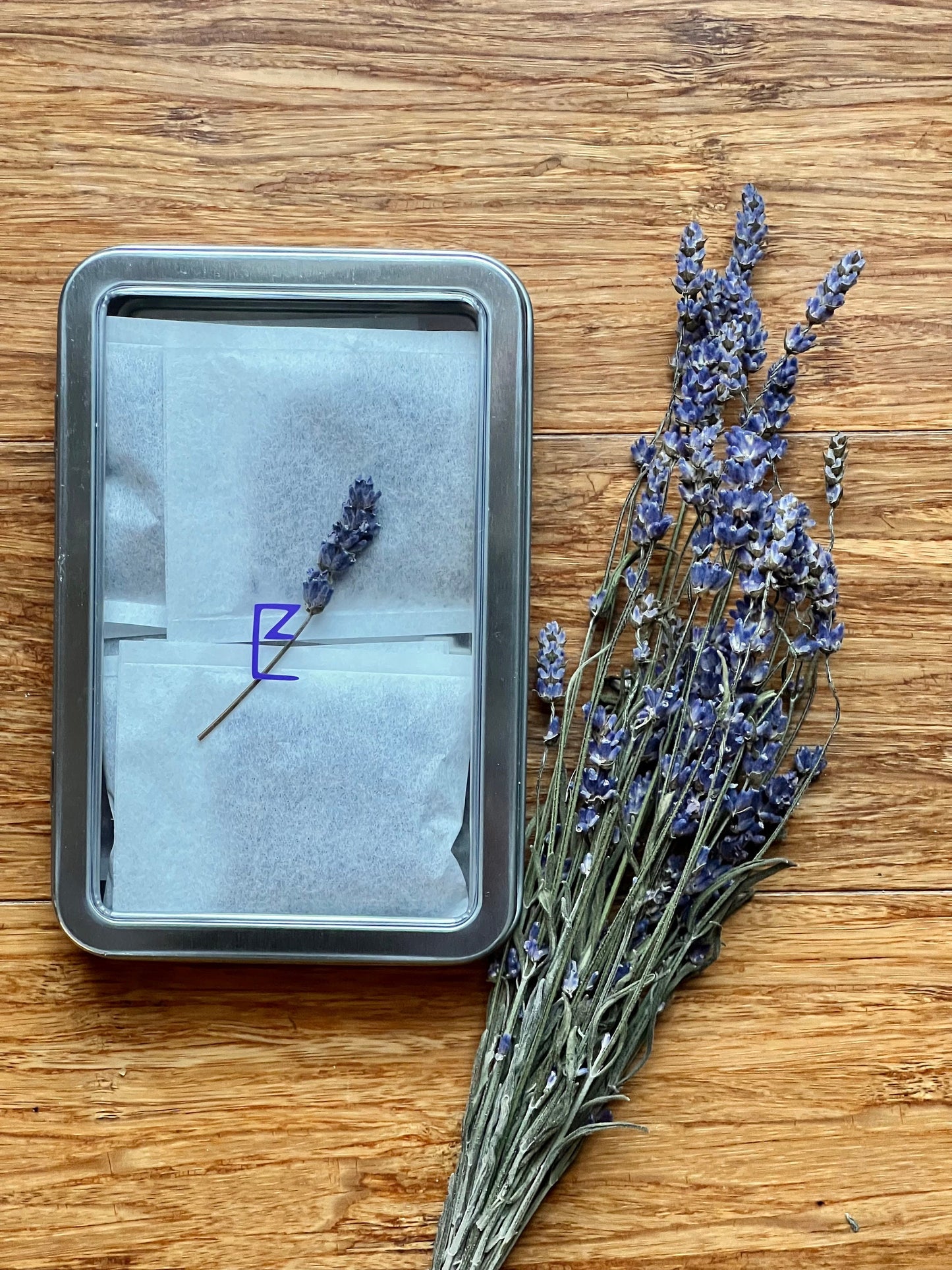 A tin box of lavender sachets placed on a wooden surface, with sprigs of dried lavender beside it, emphasizing the natural ingredients.