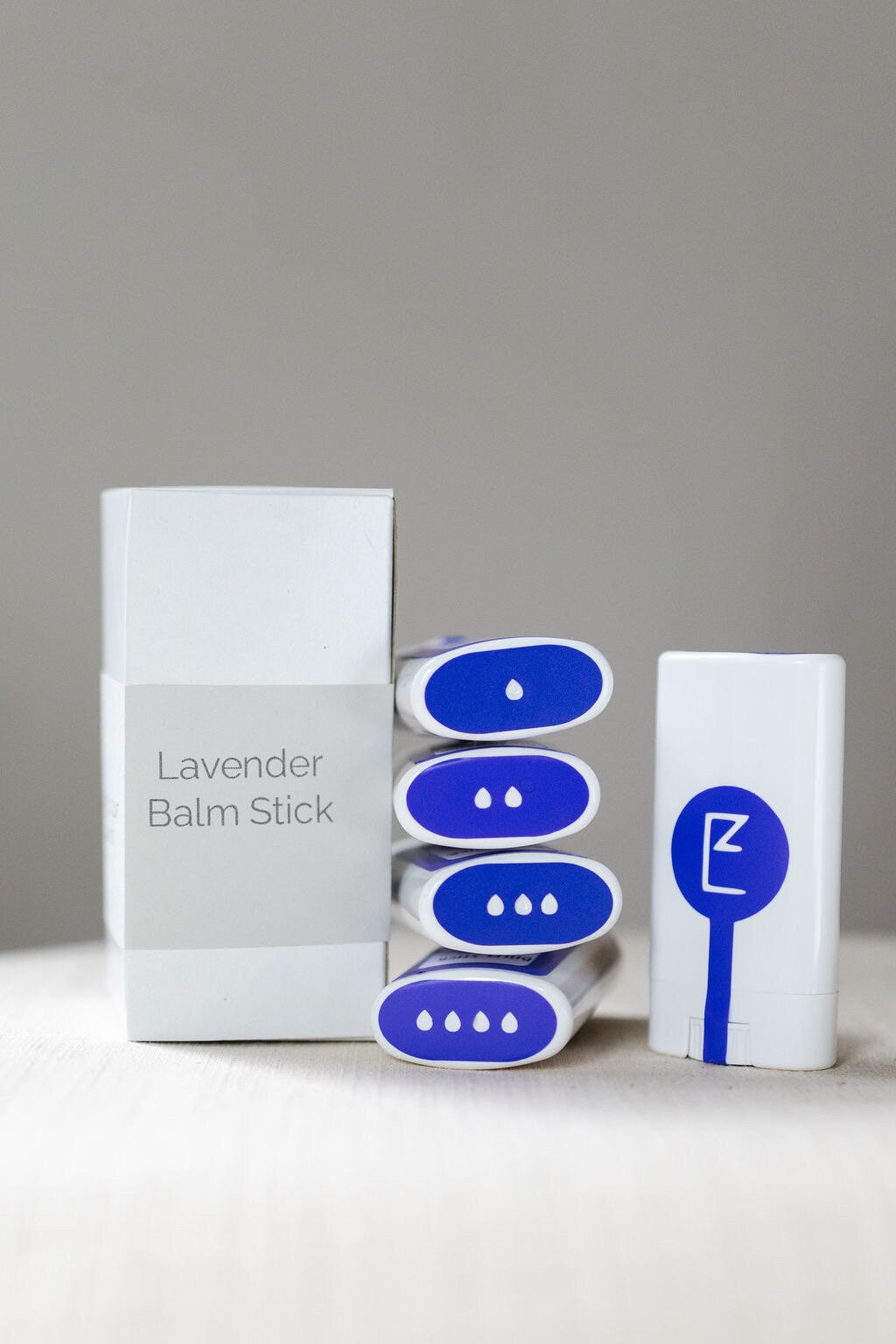 The lavender balm stick is displayed upright alongside a stack of balm sticks, and box packaging showing its compact, travel-friendly design. A solid lotion and perfume bar, made from vegan ingredients, for moisturizing dry skin, front shown.