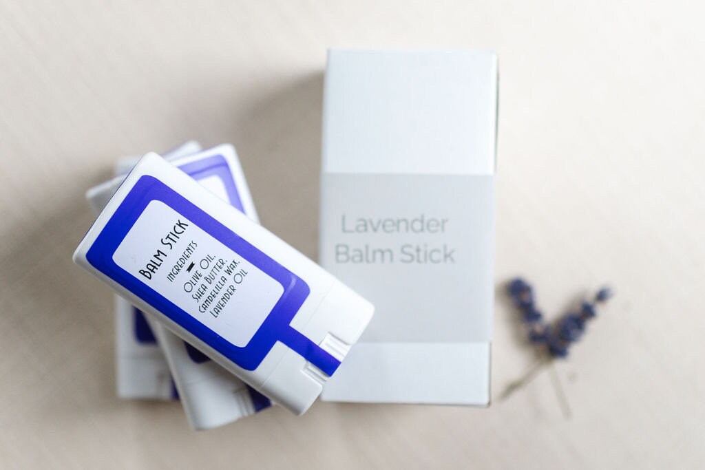 A neat flat lay of the lavender balm stick and its minimal packaging on a clean surface. The plant-based, vegan solid lotion bar is designed to hydrate and soothe dry skin while doubling as a portable lavender-scented perfume. Ingredients are shown.