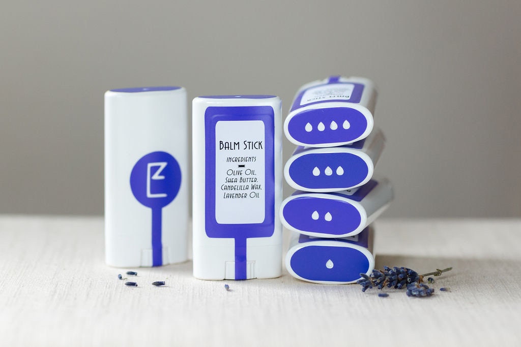 The lavender balm stick is displayed upright alongside a stack of balm sticks, showing its compact, travel-friendly design. A solid lotion and perfume bar, made from vegan ingredients, for moisturizing dry skin, four ingredients are clearly shown.