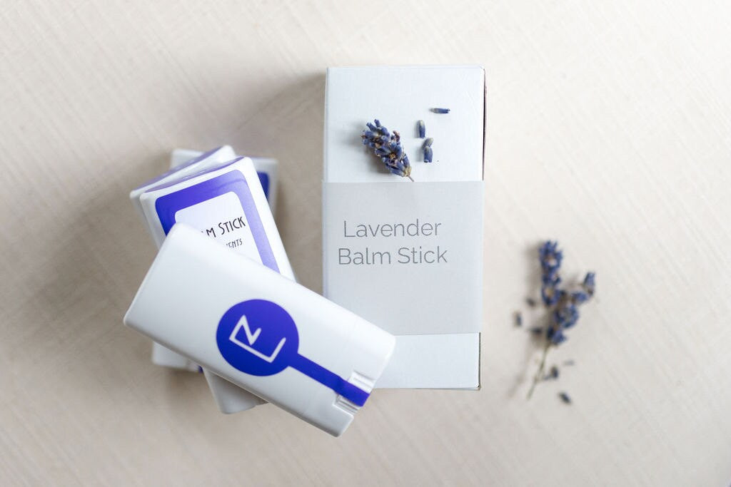 A solid lavender balm stick on a stack of balm sticks placed next to its eco-friendly packaging on a neutral surface. Showing the compact, travel-size lotion and perfume bar which is crafted with vegan ingredients and holds a calming lavender scent.