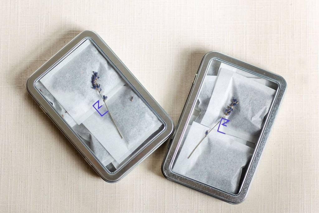 Two tin boxes of lavender sachets placed on a neutral surface, emphasizing the natural ingredients and compact size. The sachets are neatly arranged, showcasing their biodegradable packaging.