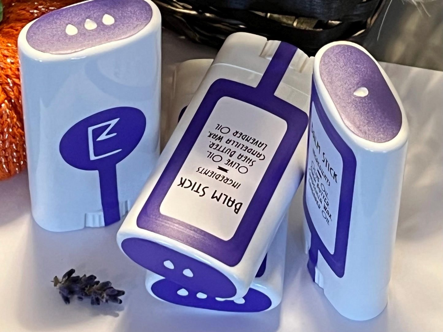 A view of the lavender balm stick and its minimal packaging in between upright travel friendly lavender balm sticks. The plant-based, vegan solid lotion bar will soothe dry skin and double as a portable lavender-scented perfume. Ingredients shown.