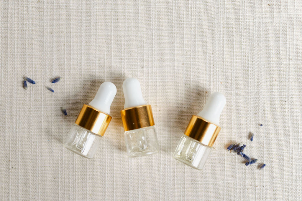 Three vials of oil laying down with lavender buds. Each vial has a white stopper, gold rim and is clear. The oil is a good color. 