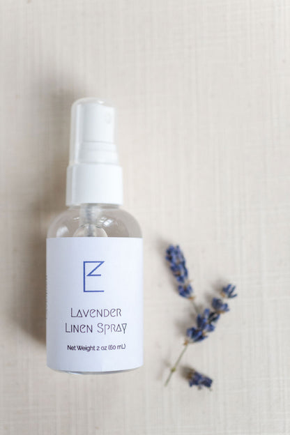 Lavender Linen Spray bottle laying down with some lavender buds shown. Bottle has a white spray nozzle with cap, clear bottle and white label. 