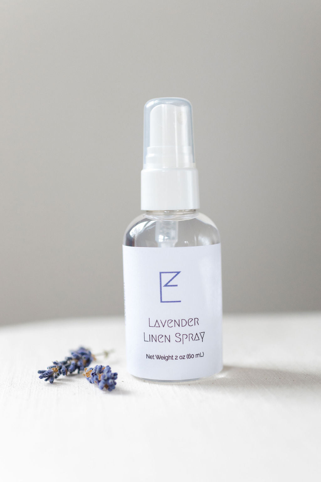 Lavender Linen Spray bottle with some lavender buds shown. Bottle has a white spray nozzle with cap, clear bottle and white label. 