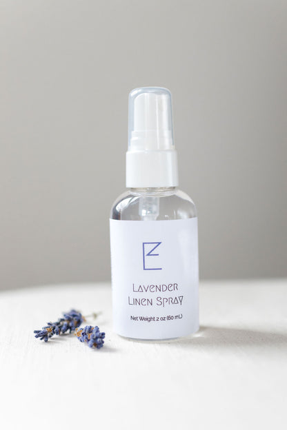 Lavender Linen Spray bottle with some lavender buds shown. Bottle has a white spray nozzle with cap, clear bottle and white label. 