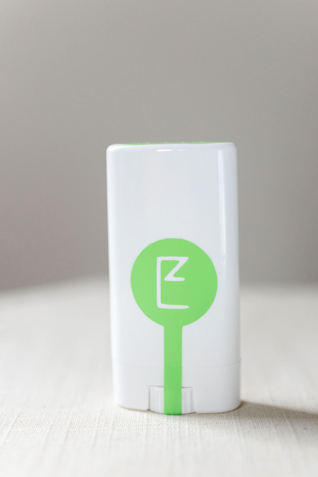 "Don't Bug Me" Balm Stick container front.  White rectangular plastic container with a thin green line form the bottom middle ending as a large green dot in the middle of the container.  Inside the dot is the E logo.