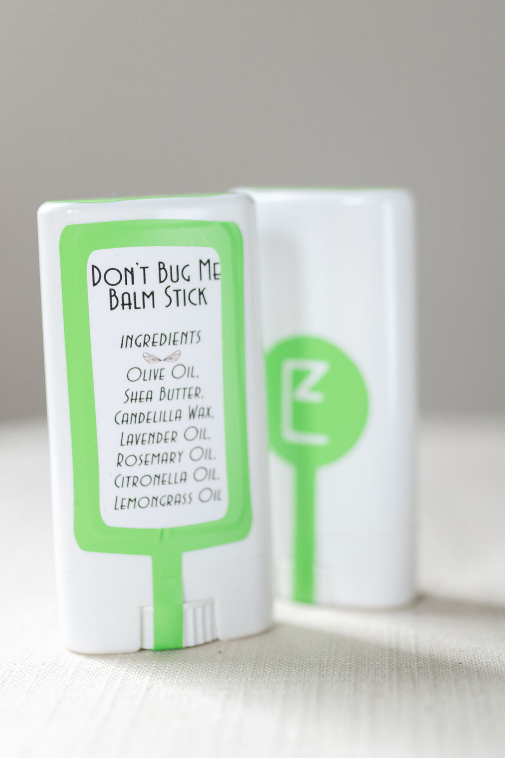 'Don't Bug Me Balm Stick' label in the foreground. A green box surrounds 'Ingredients: Olive Oil Shea Butter Candellia Wax Lavender oil Rosemary Oil Citronella Oil Lemongrass Oil'. Second Balm Stick in the background facing front. The front is white plastic with a green dot in the middle. All on a linen tablecloth.