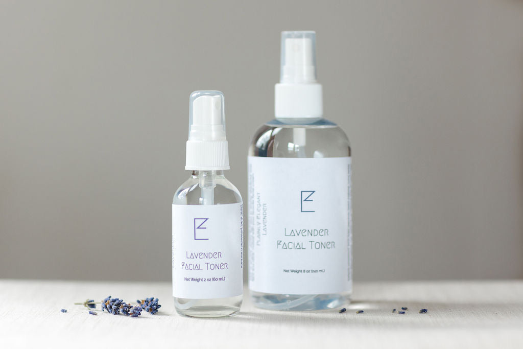 Large and small Lavender Facial Toner bottles. Both are clear bottle with white label and white spray top. The small is to the left with the large in the right. 