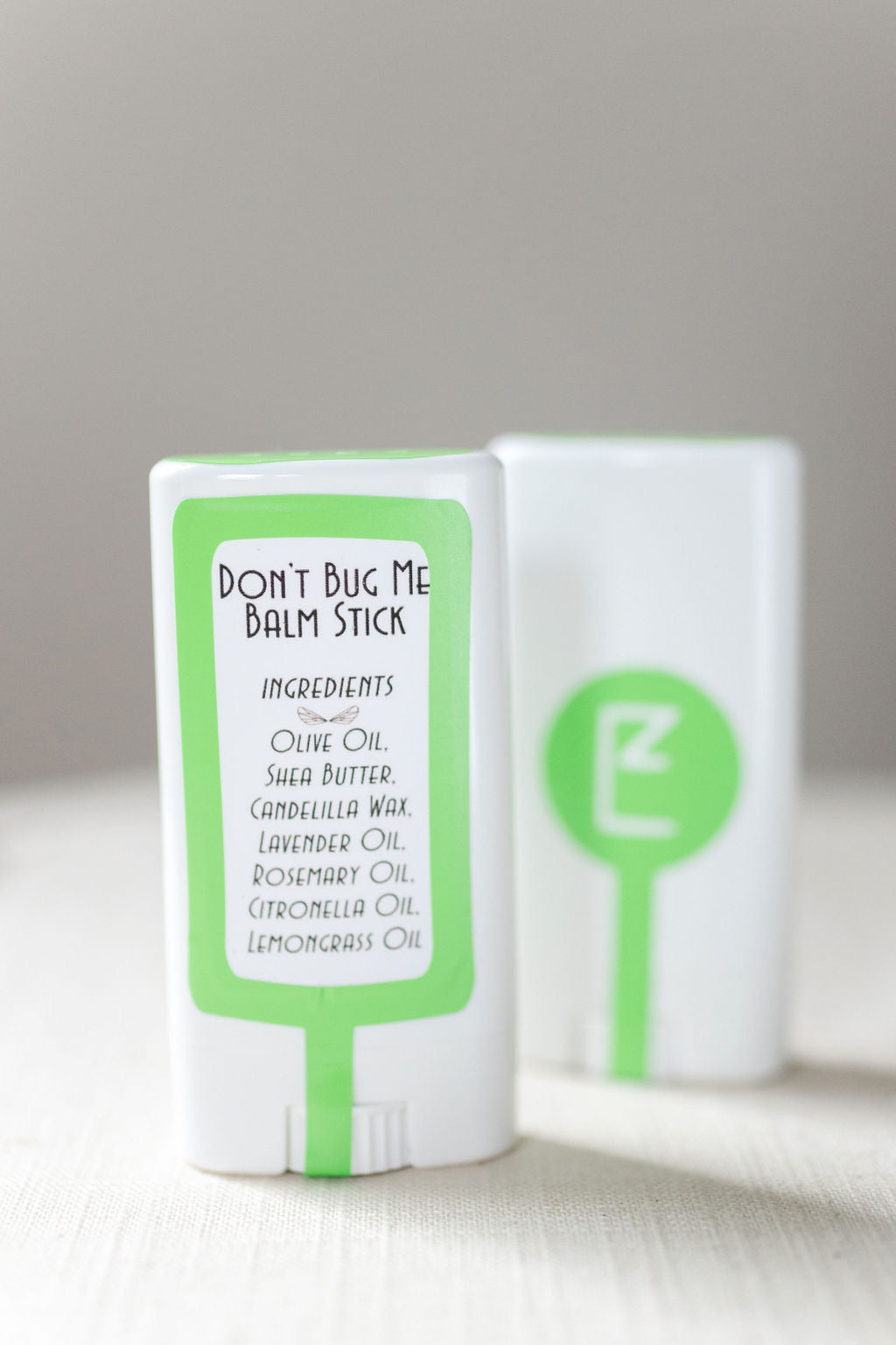 'Don't Bug Me Balm Stick' label in the foreground.  A green box surrounds 'Ingredients: Olive Oil Shea Butter Candellia Wax Lavender oil Rosemary Oil Citronella Oil Lemongrass Oil'.  Second Balm Stick in the background facing front.  The front is white plastic with a green dot in the middle.  All on a linen tablecloth.