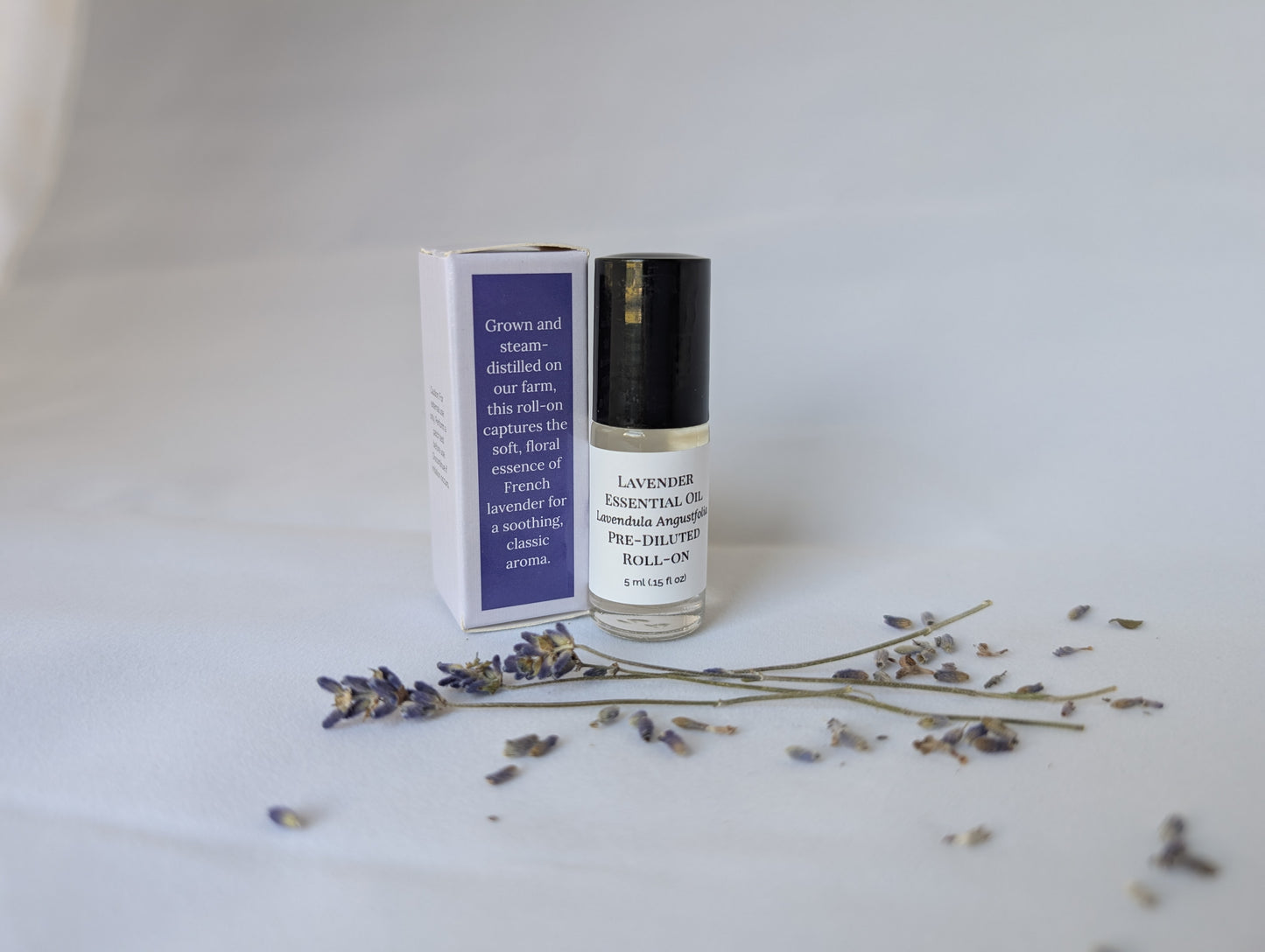 A pre-diluted lavender essential oil roll-on bottle and its white box, with dried lavender sprigs on a white surface.