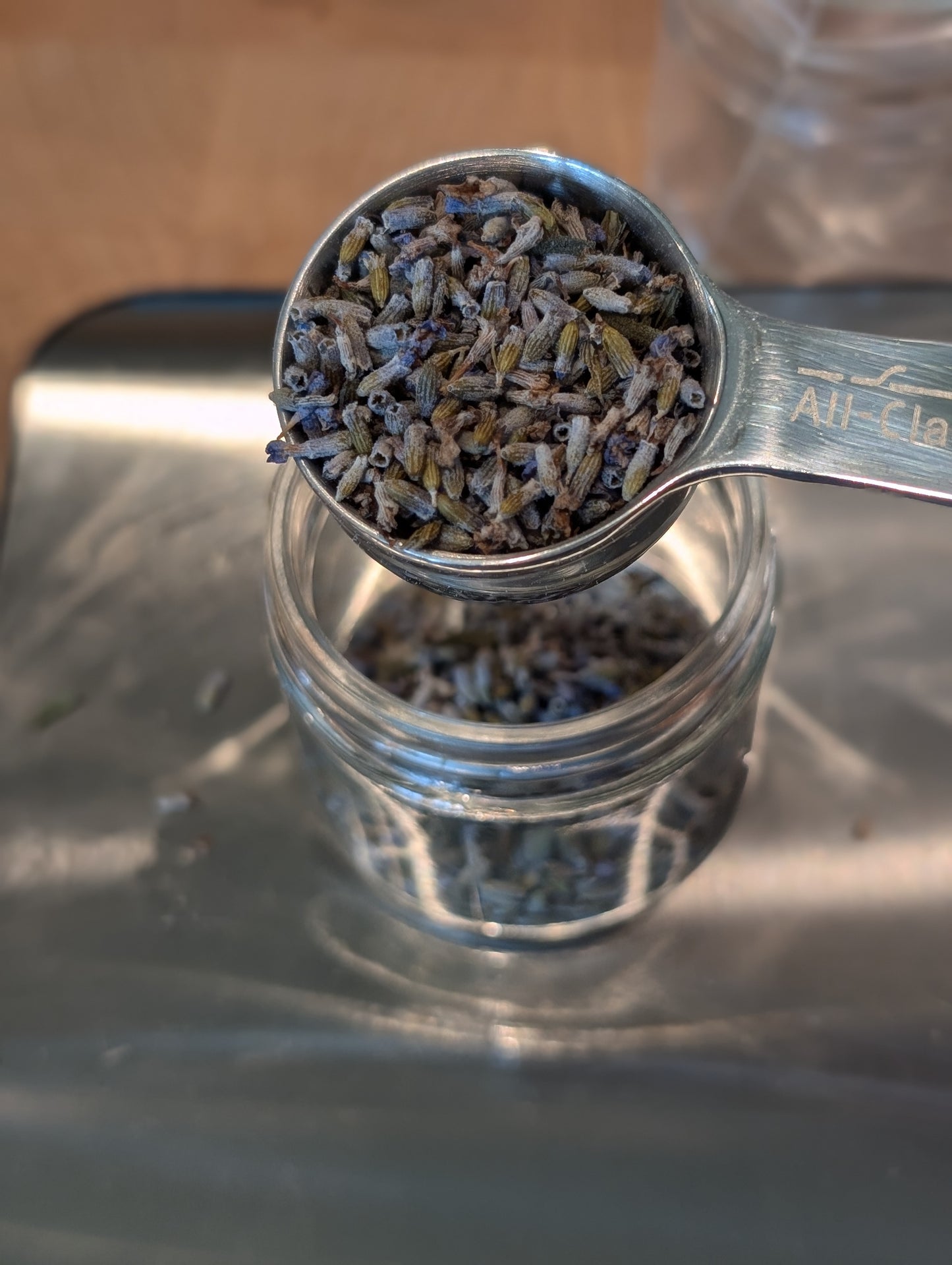 Small, dried lavender buds pour from a metal measuring spoon into a glass jar.