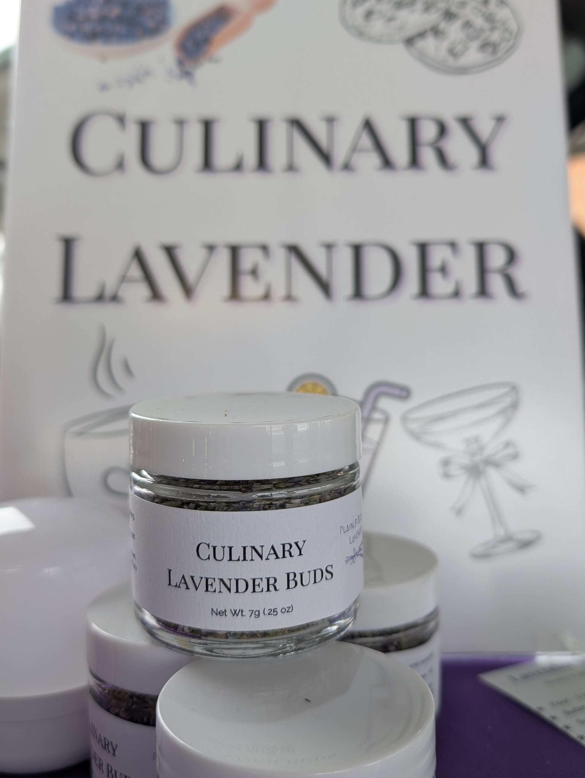 Small clear jars of Culinary Lavender Buds, 7g (.25 oz), stacked. A blurred sign in the background reads 'Culinary Lavender'.