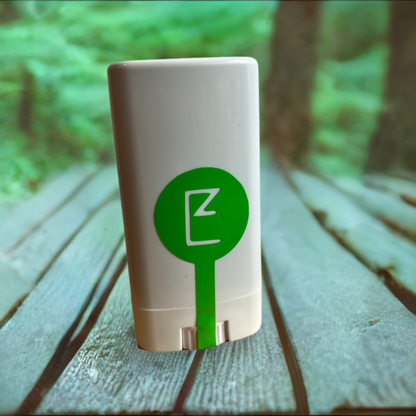 "Don't Bug Me" Balm Stick container front. White rectangular plastic container with a thin green line form the bottom middle ending as a large green dot in the middle of the container. Inside the dot is the E logo.  Background is a wood table in the forest.